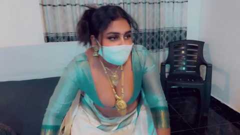 Video of a South Asian woman with medium skin tone, wearing a teal, low-cut blouse, gold necklace, and a white mask, seated in a room with a black chair and a window with white curtains.