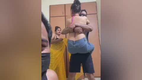 Video of a man carrying a woman in a yellow dress, with another woman in a pink top and black pants observing. Background features wooden closet doors.