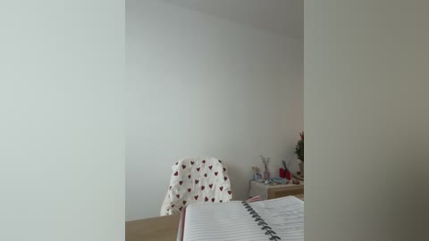 A video of a minimalist bedroom with a white wall, a wooden chair with heart-patterned fabric, a white bed with a gray sheet, and a small wooden nightstand with a vase and books.