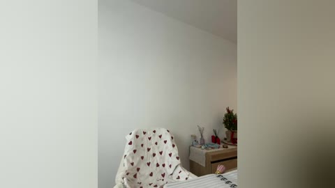 Video of a minimalist, beige-walled bedroom featuring a white chair with red heart-patterned fabric, a wooden nightstand with a red candle, and a potted plant.
