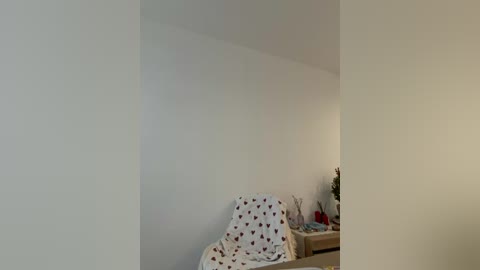 A video of a minimalist, sparsely furnished room with a white wall, a small chair covered in a white blanket with red heart patterns, and a beige table with a few decorative items.