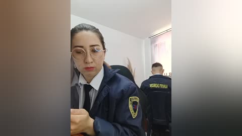Video of a young woman with light skin, wearing glasses and a navy uniform with a badge, holding a phone in a room with two police officers.