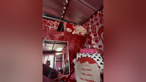 A video of a cozy, red-themed room with a plush teddy bear, a heart-shaped pillow, and a \"Polly Santa\" doll. The room features a mirrored wall, a polka-dot bedspread, and a playful, whimsical atmosphere.