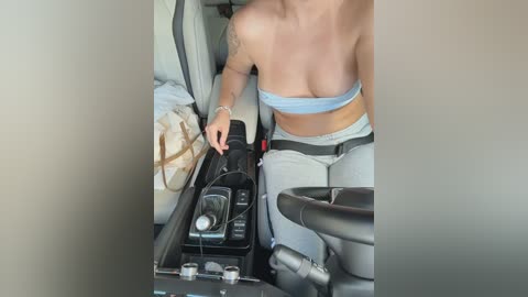 A video of a topless woman in a car, wearing a strapless light blue bandeau top and light gray pants, adjusting the car's stereo with her right hand, while her left arm rests on the car door.