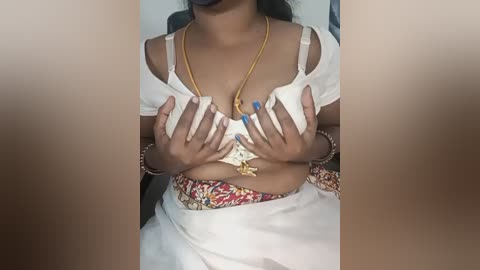 Video of a South Asian woman with medium brown skin, wearing a white saree, blue nail polish, and gold necklace, covering her breasts with her hands.
