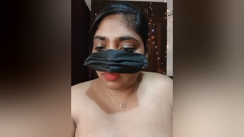 A video of a South Asian woman with medium brown skin, wearing a black mask and red lipstick, standing in a dimly lit room with a dark background.
