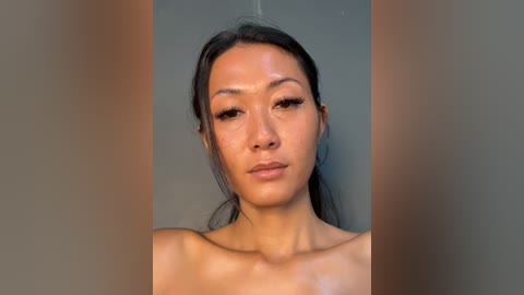 Video of a topless East Asian woman with long black hair, fair skin, and natural makeup, standing against a plain grey background.