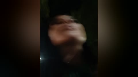 Blurry video of a person with short, dark hair, possibly female, in a dimly lit, forest-like setting, wearing dark clothing.
