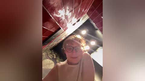 A video of a woman with curly blonde hair and glasses, wearing a beige top, standing in a dimly lit room with red tiled walls and a large mirror reflecting bright lights.