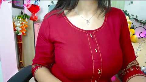 Video of a woman with medium skin tone and dark hair, wearing a red, long-sleeved blouse with gold embroidery, sitting in a cluttered room with floral arrangements, stuffed toys, and a pink chair.