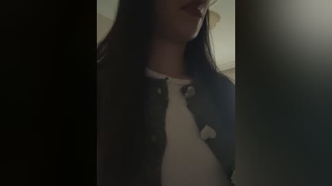 Media: Video of a young woman with long, dark hair, partially obscured by a shadow, wearing a black jacket over a white shirt. The background is dimly lit, creating a moody atmosphere.