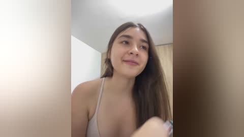 Video of a young Latina woman with long brown hair, wearing a light-colored spaghetti-strap top, smiling, in a dimly lit, beige-walled room.