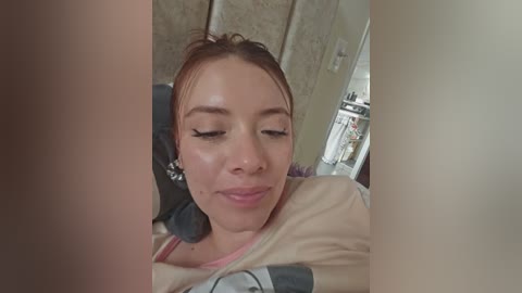 Video of a young woman with light skin, light brown hair, and closed eyes, lying on a bed with beige sheets, surrounded by blurred, soft-focus room details.