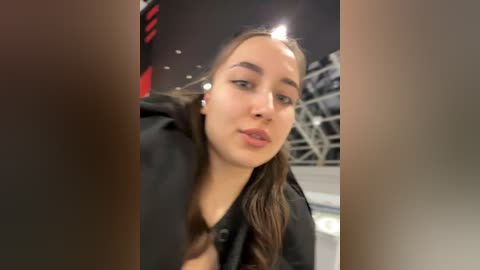 Video of a young Caucasian woman with light skin, straight brown hair, and a black jacket, making a pouty face. Background includes a blurred, modern indoor setting with metallic structures and a red and black patterned wall.