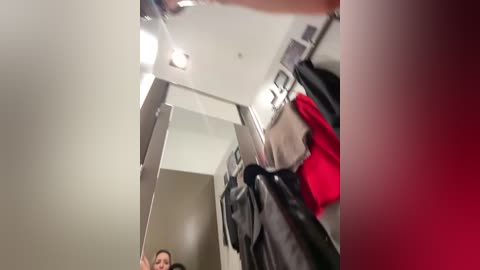 A video of a person wearing a black jacket and red shirt, standing in a modern, minimalist room with white walls and a mirror reflecting the ceiling light.