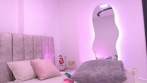 Video of a modern bedroom with a curved wall mirror, a plush grey bedspread, and a decorative Christmas tree. The room is bathed in soft purple lighting.