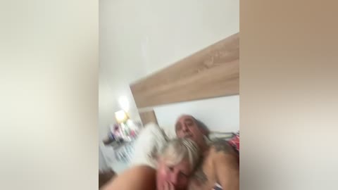 Video of a woman with blonde hair, large breasts, and tattoos, lying on a bed with a wooden headboard. She appears to be performing oral sex on a man, who is out of frame. The room has a beige color scheme.