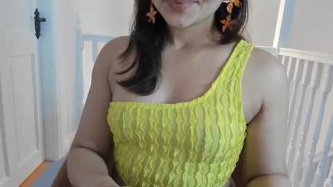Video of a smiling woman with medium skin tone and dark hair, wearing a bright yellow, ruffled one-shoulder dress and orange star earrings, in a white and blue nursery setting.