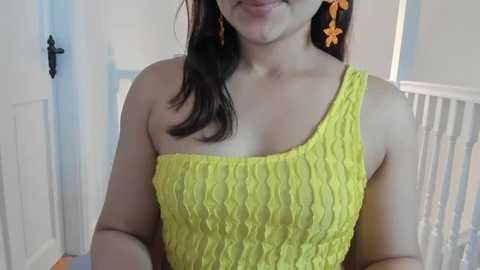 Video of a light-skinned woman with long black hair, wearing a bright yellow, textured off-shoulder dress with orange flower earrings, smiling, in a brightly lit room with white walls and a white crib in the background.