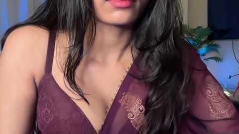 Video of a South Asian woman with long, wavy black hair, wearing a sheer, burgundy lace bralette revealing her ample cleavage. The background features a blurred indoor setting with a green plant and a flat-screen TV.