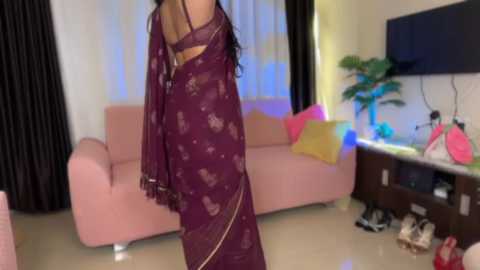 Video of a woman in a maroon saree, viewed from behind, standing in a modern living room with a pink sofa, a flat-screen TV, and shoes scattered on the floor.