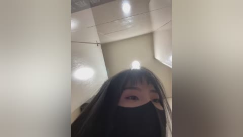 A video of an East Asian woman with long black hair, wearing a black mask, standing in a dimly lit, beige-tiled bathroom.