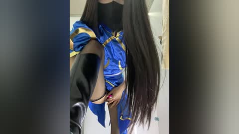 Video of a person with long, straight black hair, wearing a blue and gold outfit with a black top, standing in a bathroom with a white door and beige wall.