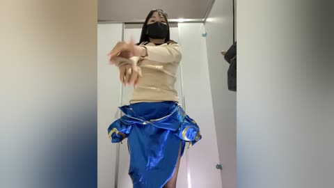 Video of a person in a beige sweater and blue skirt with a black face mask, caught mid-dance in a narrow hallway, arms flailing, with a blurred background.