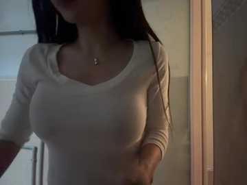 Video of a woman with long, dark hair, wearing a tight, white, long-sleeved top, standing in a dimly lit bathroom, with a visible reflection of a mirror and a tiled wall in the background.