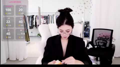 A video of a woman with dark hair in a bun, wearing a black blazer, sitting at a desk in a modern, minimalist room with a digital overlay of a fitness app.