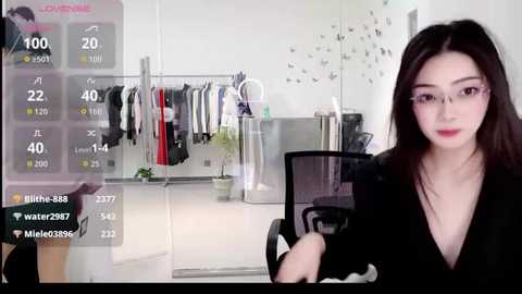 Video of an East Asian woman with straight black hair and glasses, wearing a black blouse, seated in a modern, minimalist room with a wardrobe and a plant.