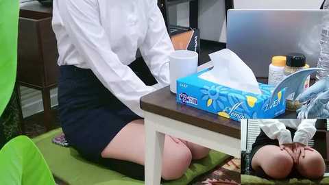 Video of a woman in a white blouse and navy skirt kneeling on a green mat, blowing her nose with a blue tissue box, in a cluttered office with a computer, coffee mug, and plants.