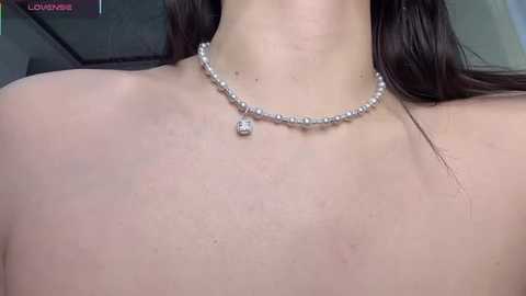 Close-up video of a fair-skinned woman with long brown hair, wearing a pearl necklace with a diamond pendant, and no visible clothing. Background is blurred, possibly indoors.