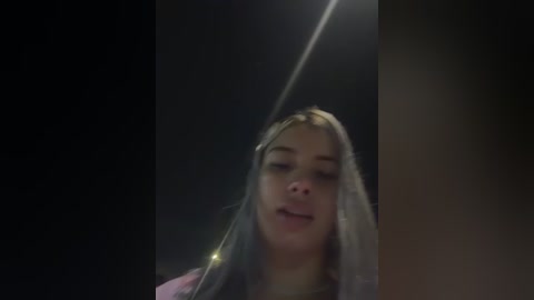 Video of a young Caucasian woman with long, straight, platinum blonde hair and fair skin, wearing a dark jacket, holding a flashlight, illuminated by a bright beam of light, against a dark background.