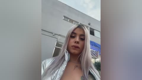 Video of a young woman with long, platinum blonde hair, wearing a partially unbuttoned white shirt, standing in front of a modern, white building with blue umbrellas in the background.