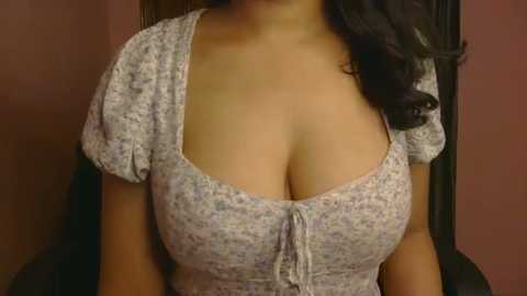 A video of a woman with medium brown skin, wearing a low-cut floral top, revealing ample cleavage and dark hair cascading over her shoulder. Background features a soft, warm-colored wall.