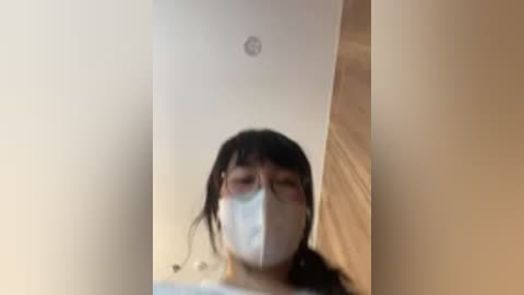 A blurred video shows a person with long black hair and a white surgical mask, standing in a room with beige walls and a wooden door, wearing a white shirt.