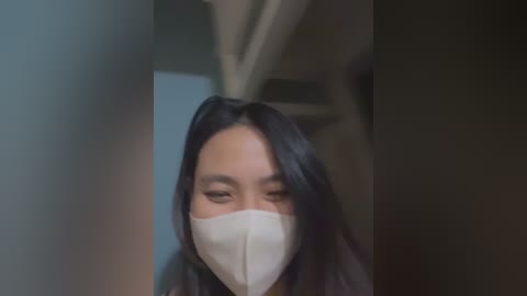 A video shows an East Asian woman with long black hair, wearing a white surgical mask and a dark top, her eyes half-closed, looking down. The background is blurred, suggesting an indoor setting.