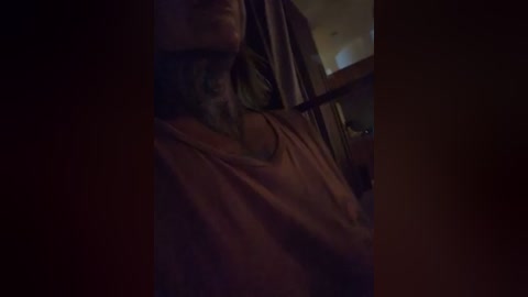 A dimly lit video captures an elderly man with a long, gray beard, wearing a dark jacket, leaning against a wall in a dimly lit room. The background is indistinct, with shadows and low lighting.