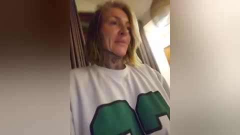 A video of a middle-aged woman with blonde hair, wearing a white t-shirt with green and black lettering. She is standing in a dimly lit room, partially obscured by a curtain.