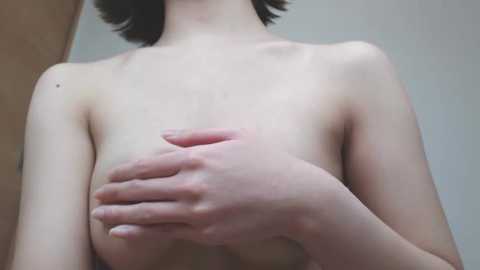 Video of a fair-skinned, topless person with small breasts, holding their right breast with their left hand. The background is plain and neutral, featuring a wooden door on the left.