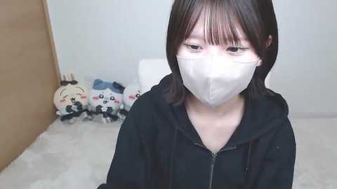 Video of an East Asian woman with straight black hair, wearing a white mask and black hoodie, sitting on a white rug in a plain room with plush toys in the background.