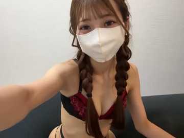 A video of an East Asian woman with long brown hair in twin braids, wearing a white face mask, a red lace bra, and beige panties, seated on a dark couch against a plain white wall.