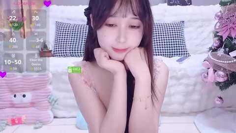 Video of an Asian woman with long black hair, resting her chin on her hands, wearing a white dress, in a cozy bedroom with a Christmas tree in the background.