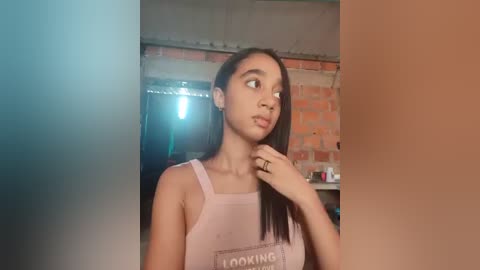 Video of a young, light-skinned woman with long, straight black hair, wearing a pink tank top with \"LOOKING\" written on it, looking contemplative in a dimly lit, rustic room with exposed brick walls.