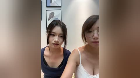 Video of two East Asian women with fair skin and dark hair. One wears a black V-neck top, the other a white lace camisole. Background features framed artwork and a hallway.