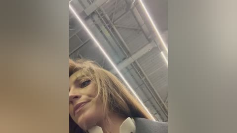 A video of a smiling woman with light skin and long, straight brown hair, wearing a white shirt, sitting in an industrial space with exposed pipes and a metal ceiling.