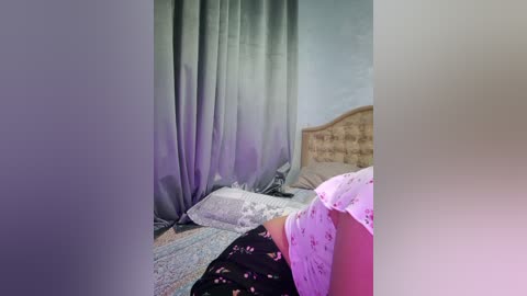 Video of a bedroom with a light gray curtain, a bed with a floral-patterned pillow, and a headboard. The background is blurred, with purple lighting creating a soft, dreamy ambiance.