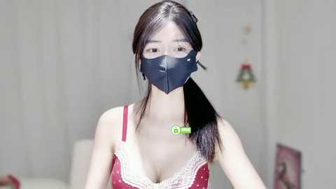 Media: A video of an East Asian woman with long black hair, wearing a red lace-trimmed bra and a black face mask, standing in a softly lit, minimalist bedroom.