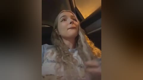 A video of a young woman with light skin and long, wavy blonde hair, wearing a white blouse, captured in a dimly lit car interior. She looks upward, lips slightly parted, and has a serene expression.